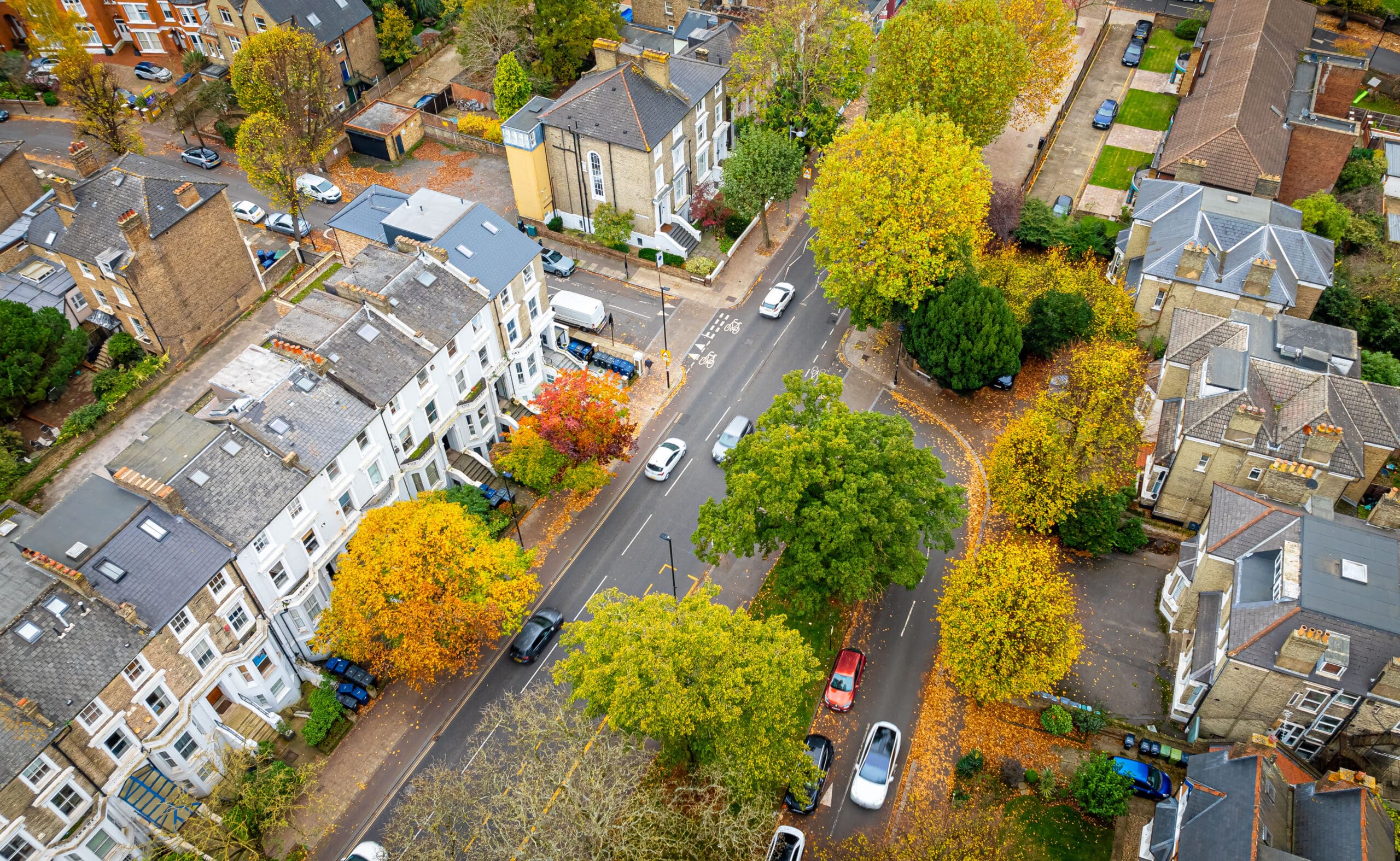 Aerial view of a tree-lined street in a residential neighborhood, with cars parked along the road and autumn leaves scattered about, surrounded by houses and buildings—many featuring modern central heating installations.