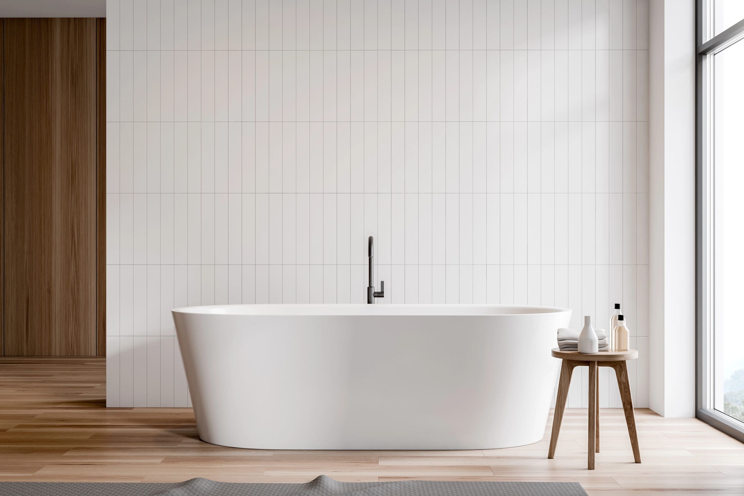 A modern bathroom with a freestanding white bathtub, sleek black faucet, and a small wooden stool for toiletries. Light wood flooring, white tile walls, and a large window complete the look—ideal after expert boiler servicing keeps things cozy year-round.